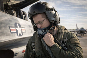 Person in military gear with a helmet and oxygen mask wearing eyeglasses in front of an aircraft on a runway.
