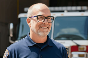 Man wearing glasses and a blue shirt with a patch, standing in front of a fire truck.